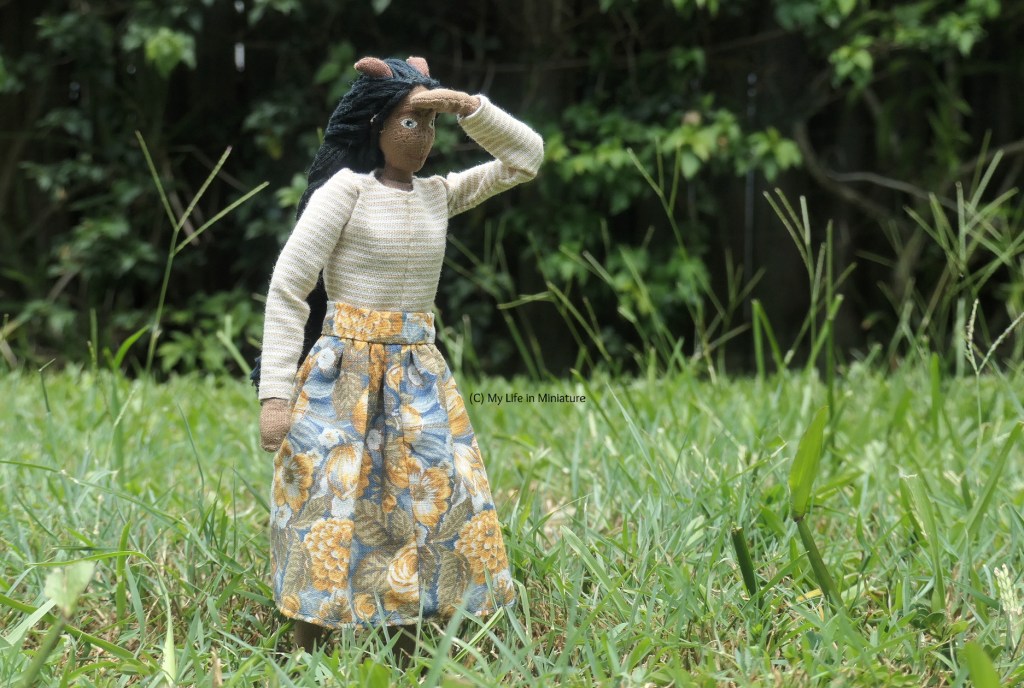 Petra stands outside in grass, with a dark hedge in the background. She wears the blue-and-orange skirt with the beige top and brown boots. She's looking to the right of the camera, one hand shading her eyes and the other by her side. 