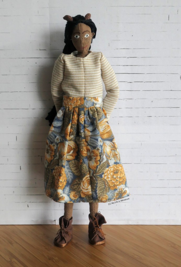 Petra stands in front of a white brick background, smiling at the camera. She has her hands behind her back, and is wearing the same blue-and-orange skirt with a beige-and-white striped top and brown boots. 