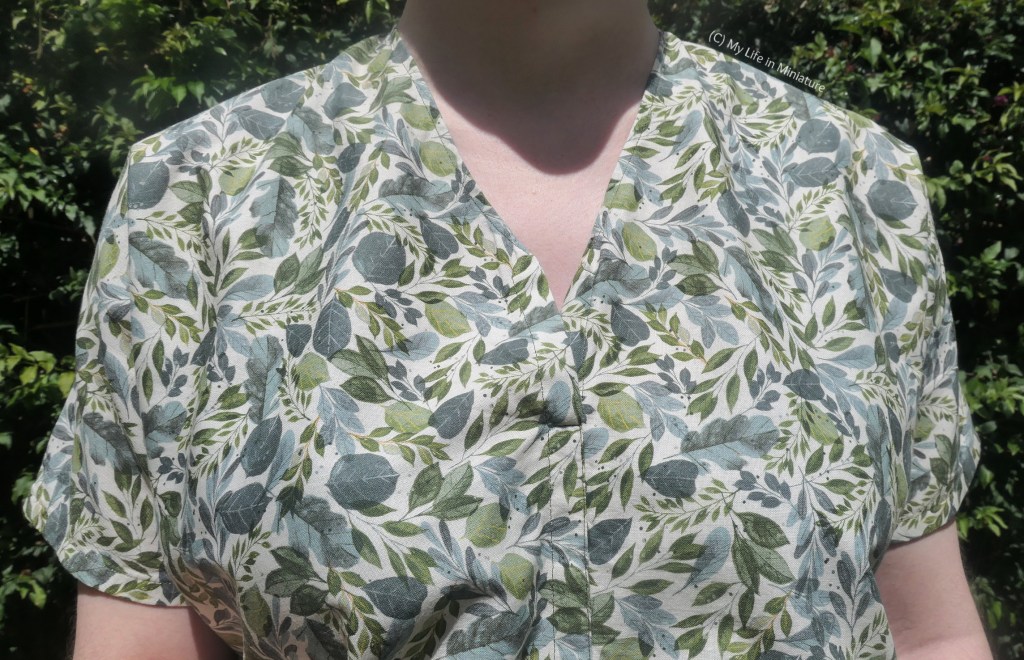 Close-up of the shirt. The pattern is a variety of different shaped leaves, in various shades of green, on a white background. The centre front is sewn shut most of the way up to the V-neckline, and the sleeves are short. 