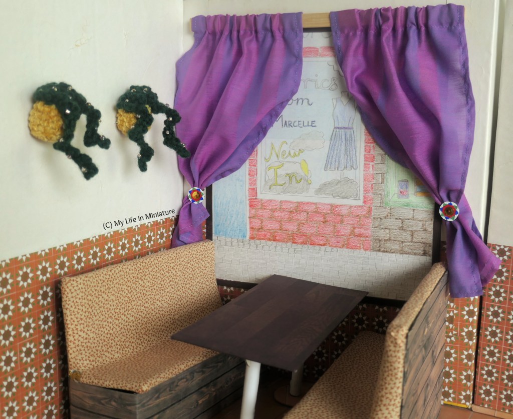 The booth under the front window at Tea at Yaz's is photographed. The window looks out onto the street and the shops opposite, with purple curtains held back by wall-mounted curtain holders. On the wall beside the window are two more hanging baskets planted with floral vines. The table and booths are empty.