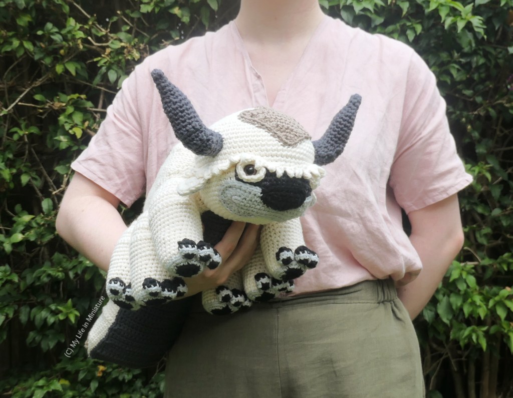 The author stands in front of a hedge, wearing a pale pink shirt and khaki pants. She's holding the toy under one arm, with his head pointing towards the camera. His eyes are surrounded by white, and he has a black nose and grey jaw.
