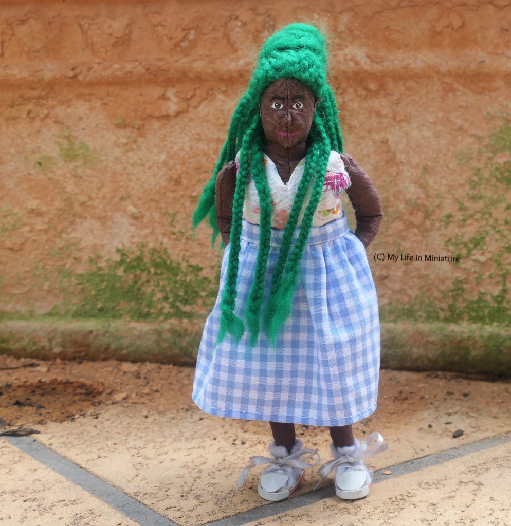 Hazel stands outside, with a mossy terracotta pot as her background. She's wearing the pale blue/white checked skirt with a white floral top and white shoes. She smiles at the camera with her hands in the skirt's pockets. Some of her braids are pulled forward over her shoulders. 