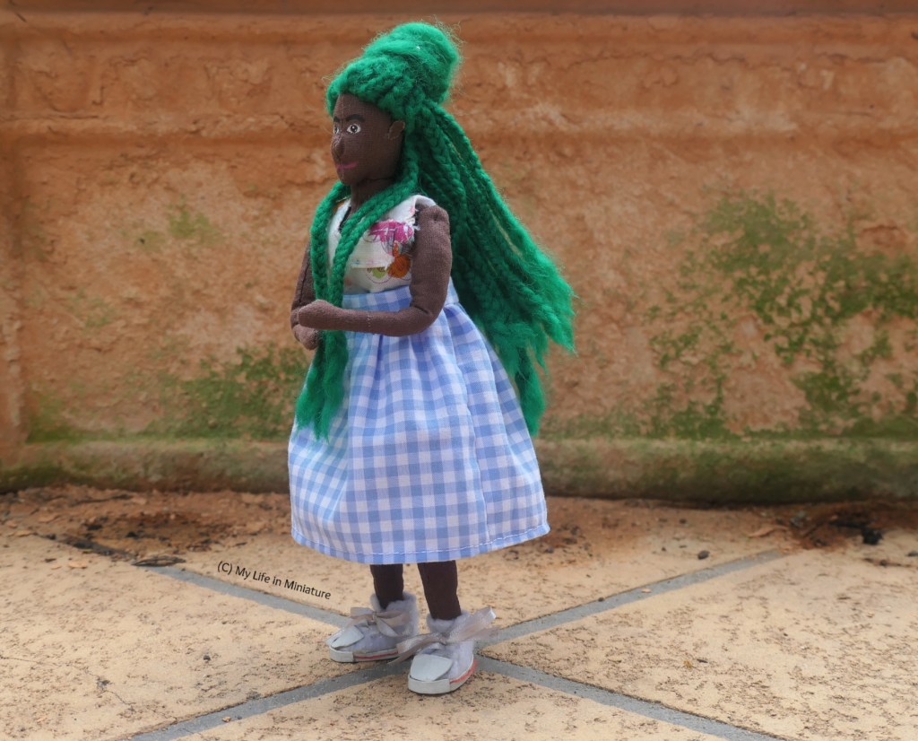 Hazel stands in front of the mossy terracotta pot, wearing the same pale blue checked skirt and floral top as before. She's twirling in place, making her hair and her skirt swing out with momentum.