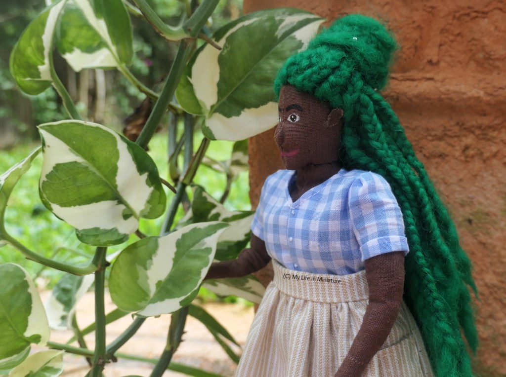 Hazel stands in front of a terracotta pot, near a cascading vine with white and green leaves. She wears the top of the pale blue/white check dress with a cream/white striped skirt. She's photographed from head to thighs, cupping one of the leaves of the vine in her hand.