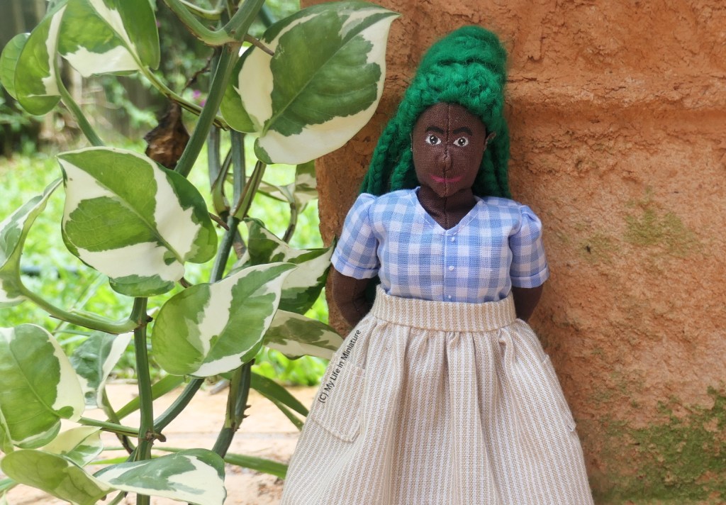 Hazel leans back against the terracotta pot, smiling at the camera with her hands behind her back. She's wearing the blue checked top and cream skirt from before. Next to her is the green and white cascading vine.