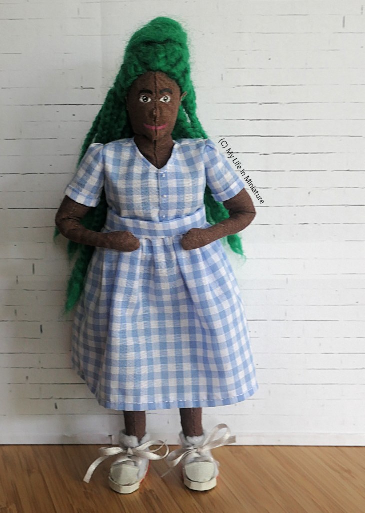 Hazel stands in front of a white brick bcakground. She wears a pale blue/white checked top with short sleeves, and a matching gathered skirt. She's smoothing down the gathers while smiling at the camera. 