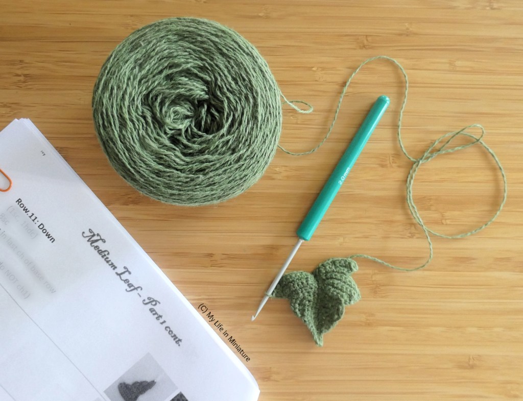 A ball of green yarn, a crochet hook, and a paper pattern are laid flat on a wood surface. The hool is connected to the yarn and to a small crocheted ivy leaf shape. The pattern says 'Medium Leaf - Part 1 cont.' across the top of the page. 
