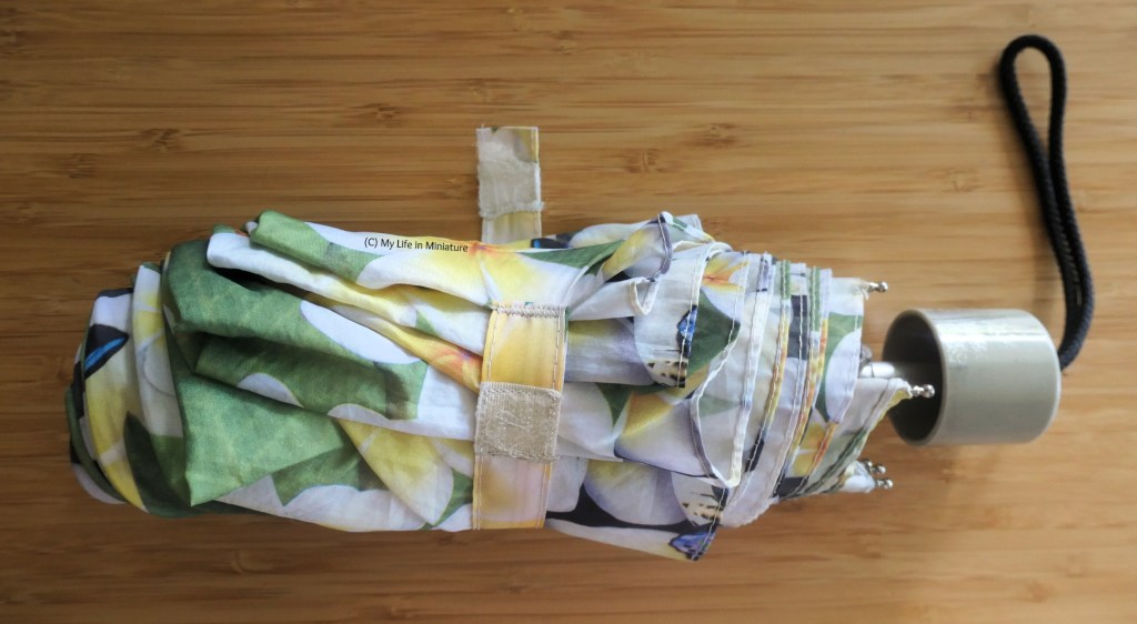 A rolled-up umbrella in shades of white, yellow, and green sits on a wood background. The Velcro strap to close it is unfastened, and one end is flat on the surface beneath.