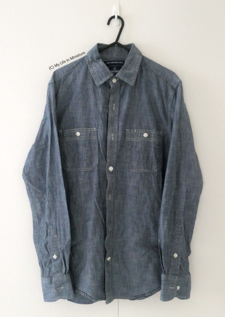 A chambray blue button-down shirt hangs from a hanger on a hook on a white background. It has white buttons, and two pockets on the chest. 