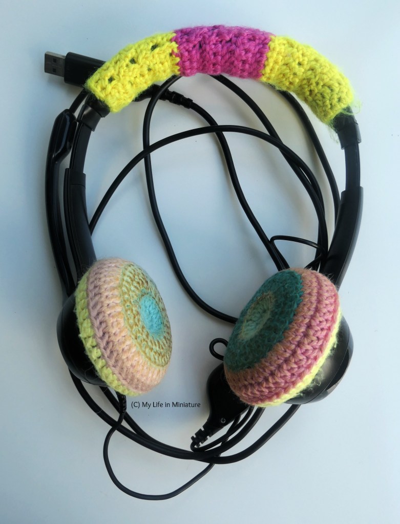 The author's over-head headphones are laid flat on a white surface. They are black, and have colourful crocheted covers over the cushioned earmuffs and overhead bit. 