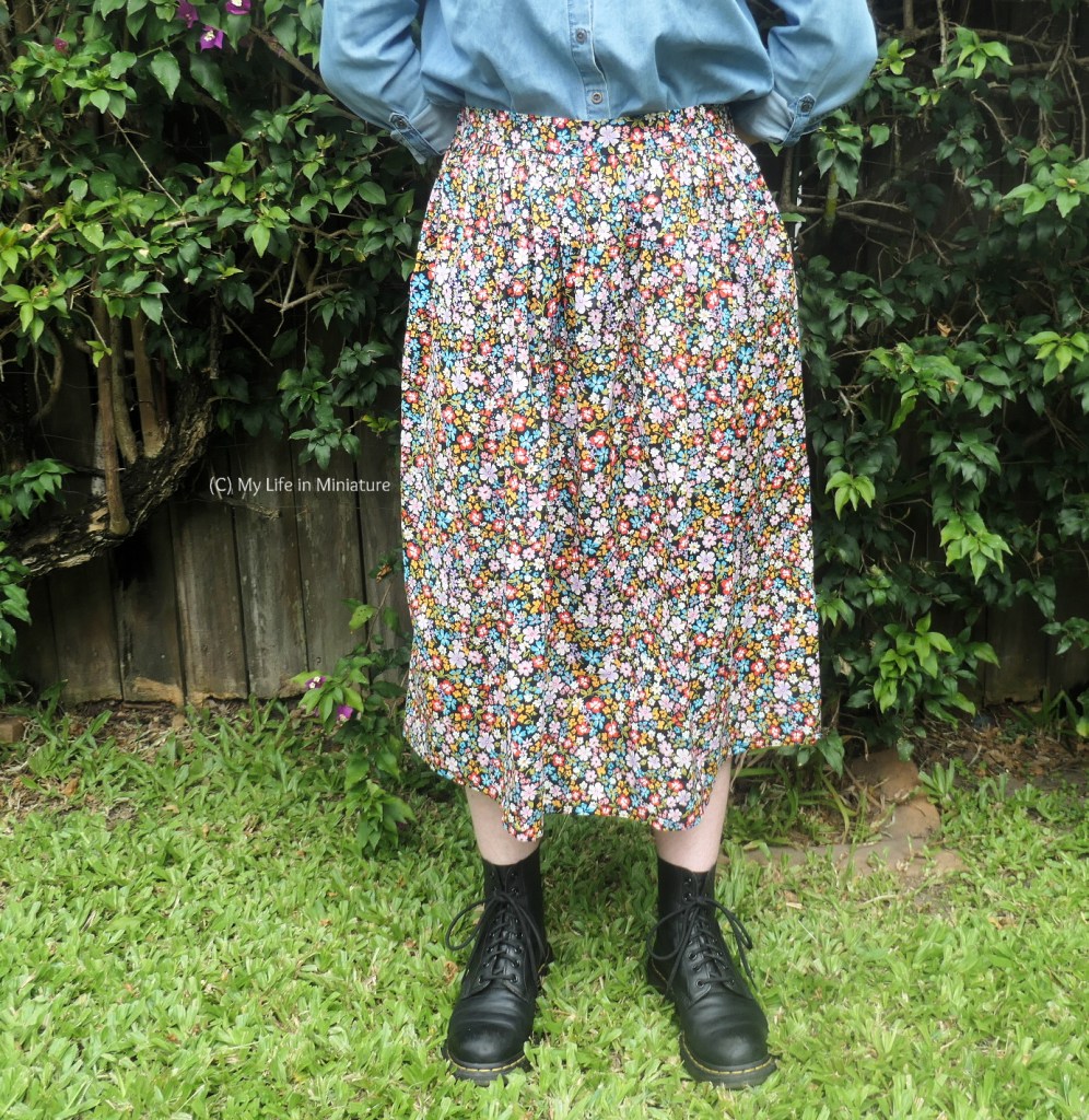 The author stands in front of a hedge, photographed from waist to the ground. She wears a floral gathered skirt with a denim blue shirt and black boots. She has her arms behind her back and faces the camera. 