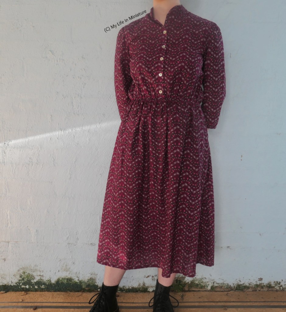 The author stands against a pale blue wall, photographed from neck to ankles. She wears a dark pink, high-collared, elastic-waisted midi dress with black boots. Her hands are tucked behind her back. 