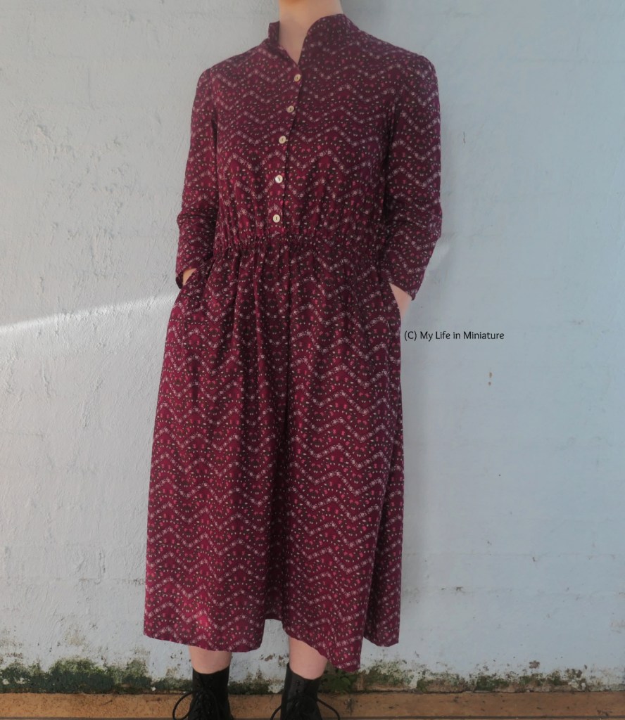 The author stands against a pale blue wall, photographed from neck to ankles. She's wearing the dark pink dress with black boots, and has her hands in her pockets.