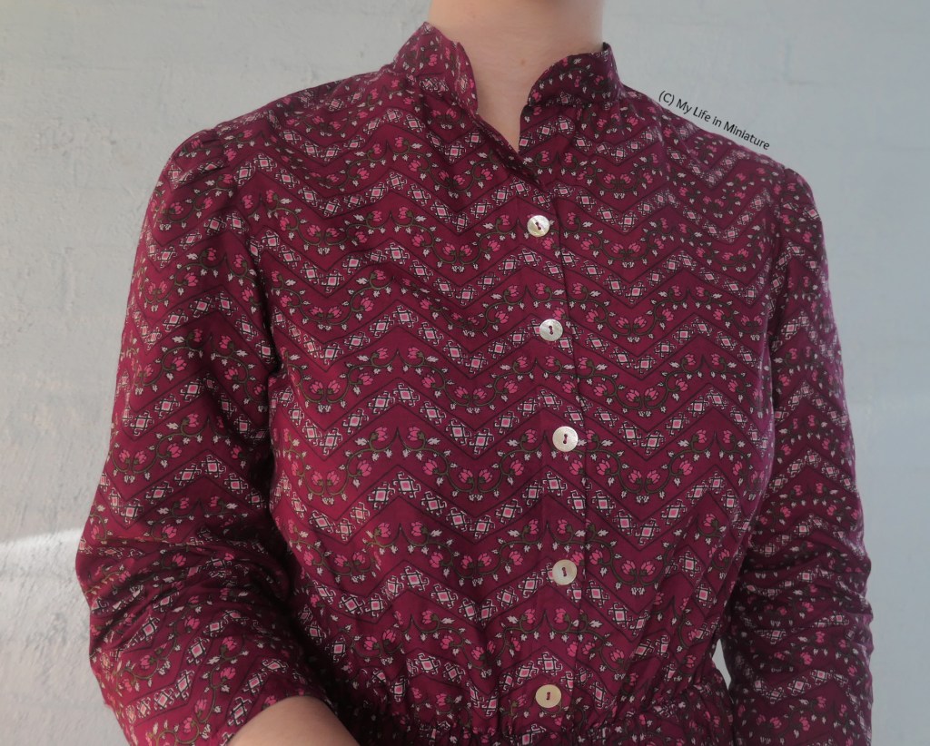 Close-up of the bodice of the dress. The fabric has horizontal zigzags filled with curly shapes, and in-between zigzags are rose vines. The buttons on the bodice are pearlescent, and the collar is a high Nehru collar, with curved corners. 