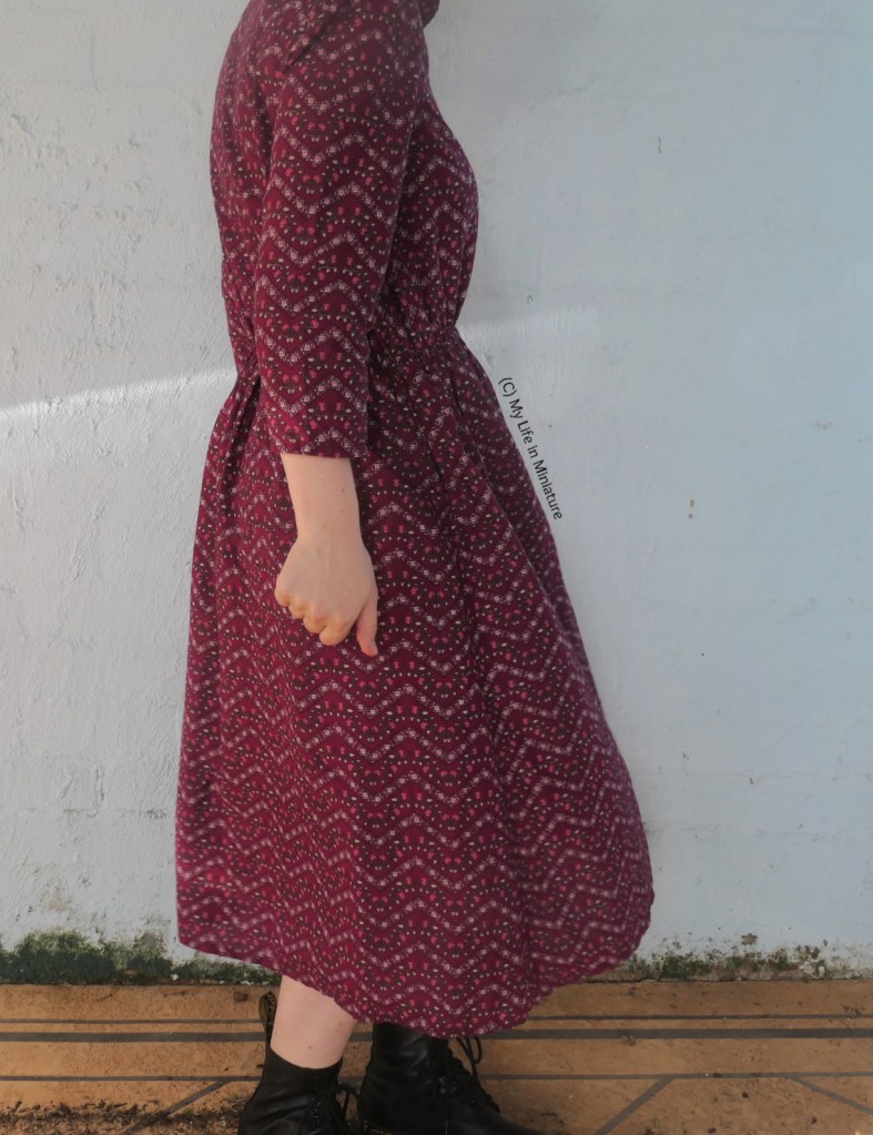 The author stands against a pale blue wall, photographed from neck to ankles. She is twirling in the dark pink dress, the skirt flaring out a little bit. 