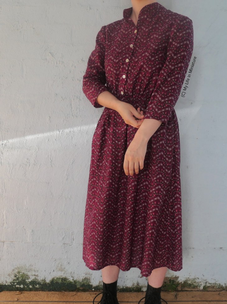 The author stands in front of a pLe blue wall, photographed from neck to ankles. She wears a dark pink midi-length dress, with small floral zigzags all over. The dress buttons up the front bodice, and is three-quarter sleeved. 