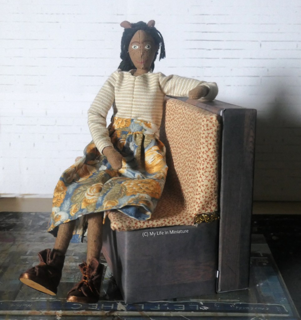Petra sits on one of the bench seats from Tea at Yaz's, which is in front of a white brick background. The back of the bench seat has been extended, so there is a small shelf behind it. Petra has one arm up on this shelf, smiling at the camera and wearing a blue and orange floral skirt with a stripy cream shirt and brown boots.