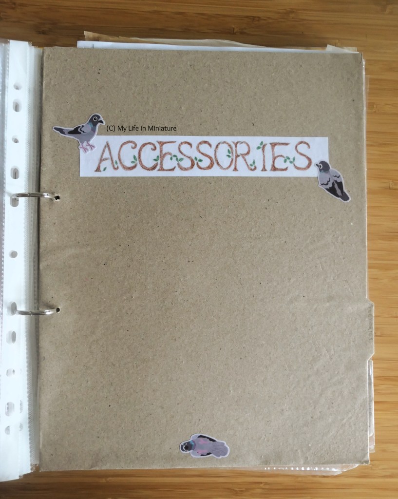 The completed section divider for "Accessories", in the binder. It looks very similar to the first divider - brown paper, label in the same font, tab near the bottom right this time. There are three stickers in the same places as before, this time of rock dove pigeons.