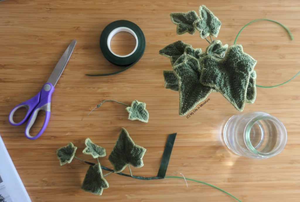 A crocheted plant in mid-assembly on a wood surface, photographed from above. There is a curl of green wire, a glass jar with crocheted leaves in, another jar with water in, a roll of florists' tape, a pair of scissors, a loose leaf, a half-assembled vine, and the instructions for the project in the bottom left corner of the image.