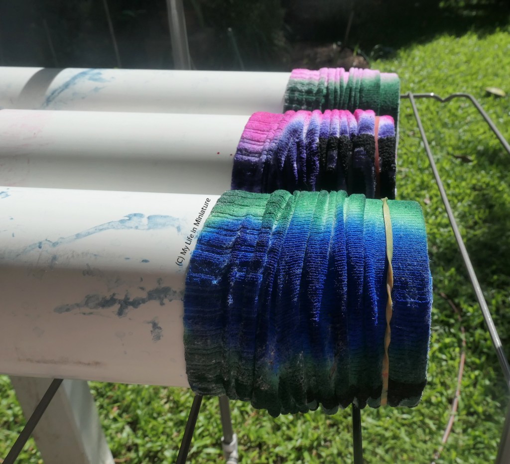 The sock-covered ends of three pipes sit on a flat, sturdy, wire washing line, with grass in the background. From the foreground, the socks are dyed green and blue, pink and purple, and pink and green. The sun is shining on the socks and the grass.