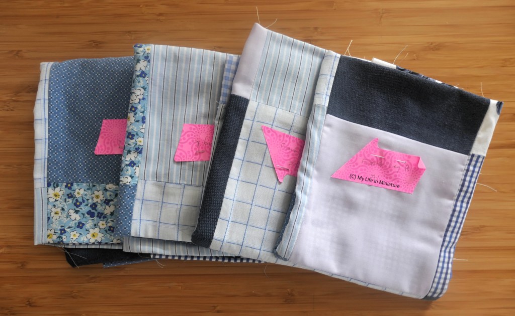 Four folded patchwork fabrics are laid out in a fan on a wood surface, photographed from above. Each has a pink sticky note pinned to the front, with indiscernable writing on. They are all in shades of blue.