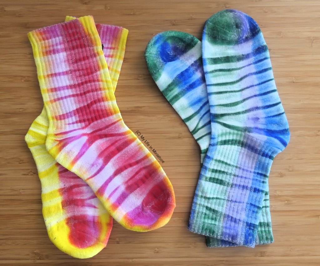 Two pairs of long crew socks are laid flat on a wood surface. One pair is dyed pink, red, and yellow, and the other is blue, green, and purple. These are also striped horizontally with irregular lighter and darker stripes. 
