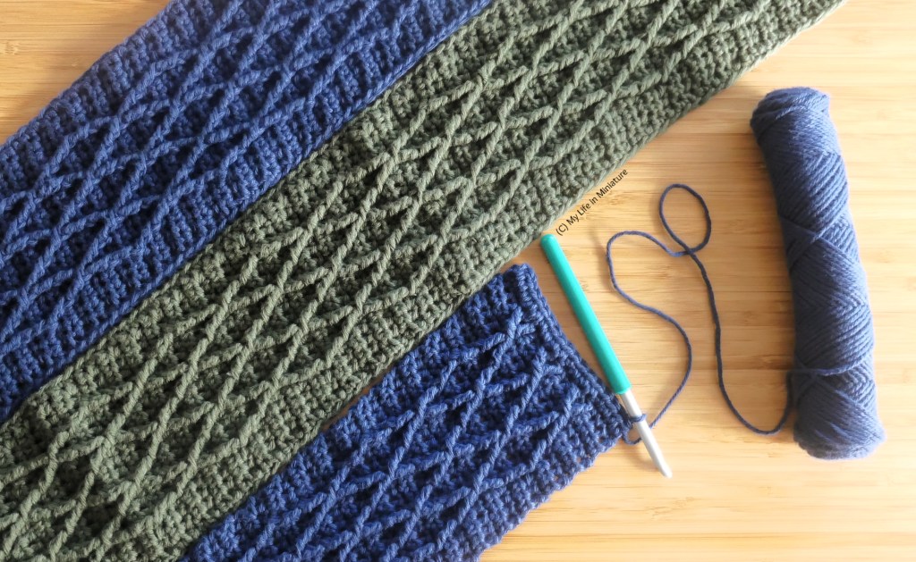 The three crochet strips are laid flat next to each other on a wood surface, photographed from above. The strip in progress is positioned halfway along the green strip, with the hook and working yarn beside it. The other navy strip is on the other side of the green strip.
