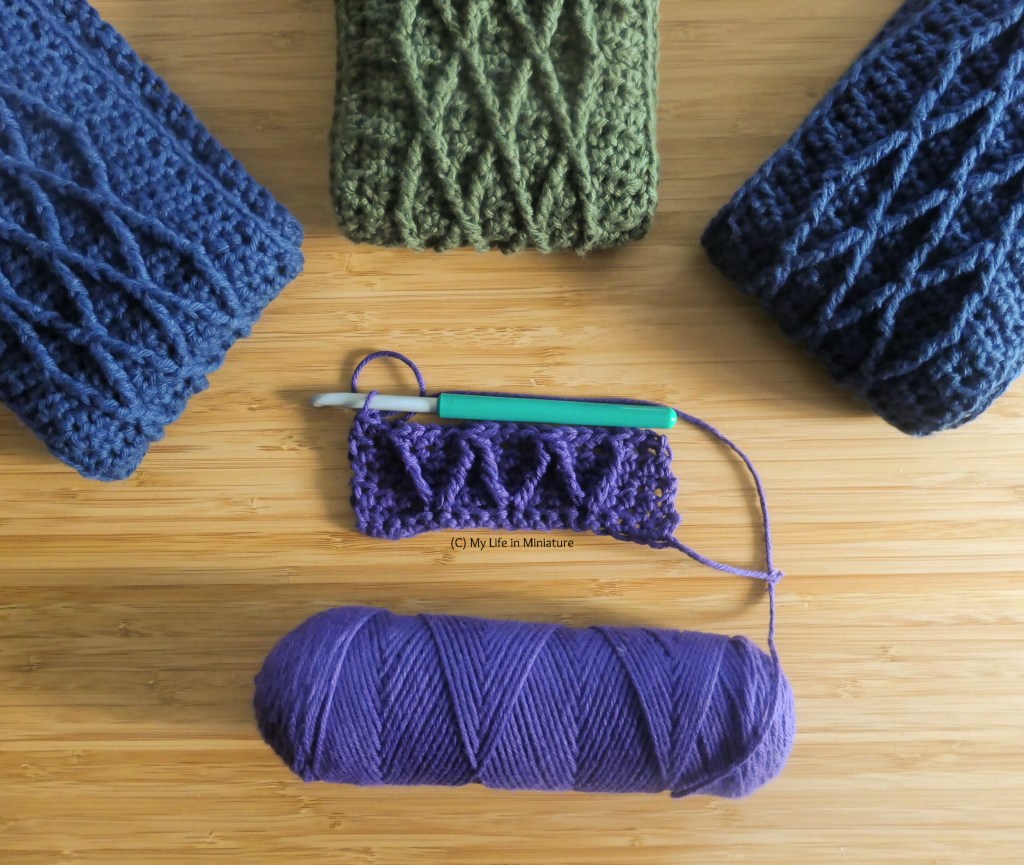 Four pieces of crochet are laid out flat on a wood surface. All are cabled; three of them are long strips folded up and arranged in a fan at the top of the image. Two of these are navy and the centre one is green. The bottom one is very small, and purple, and has the hook and ball of yarn attached. 