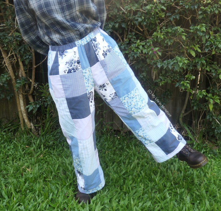 The author stands in front of a hedge, photographed from waist to ground. She's wearing a flannel shirt with a pair of patchworked, quilted pants, and brown boots. The patches are patterned and plain, in shades of blue and purple. She stands on one leg with her arms behind her back.