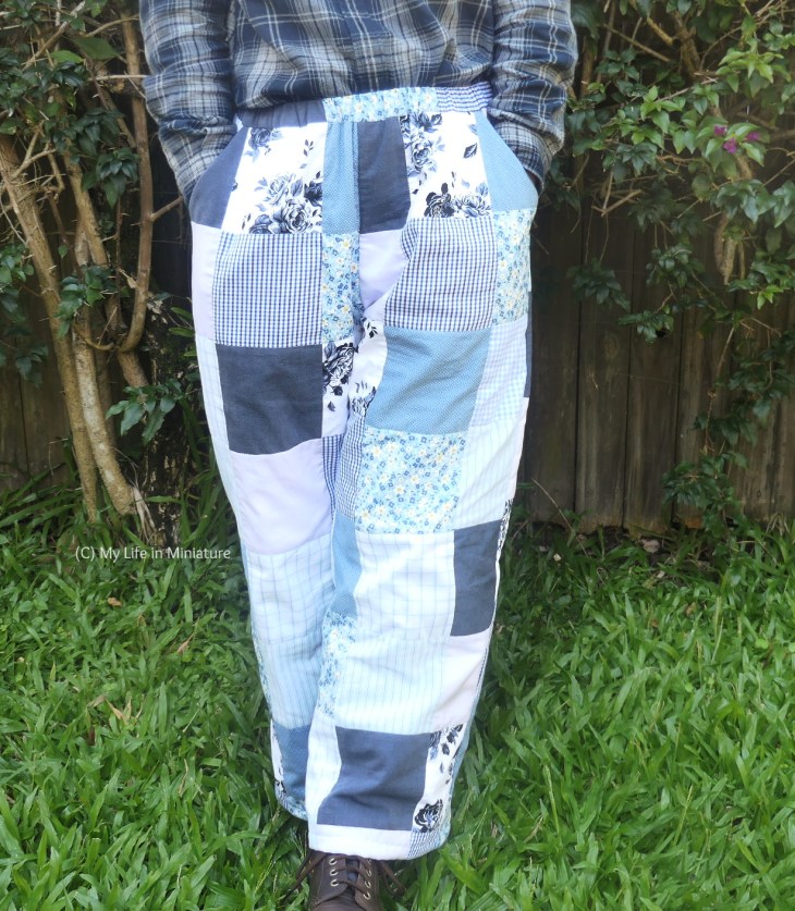 The author stands in front of a hedge, photographed from waist to ground. She wears a pair of patchworked, quilted, elastic-waisted pants with a flannel shirt tucked in and brown boots. Her hands are in the pockets of the pants, whose fabrics are an assortment of blues in various shades and prints.