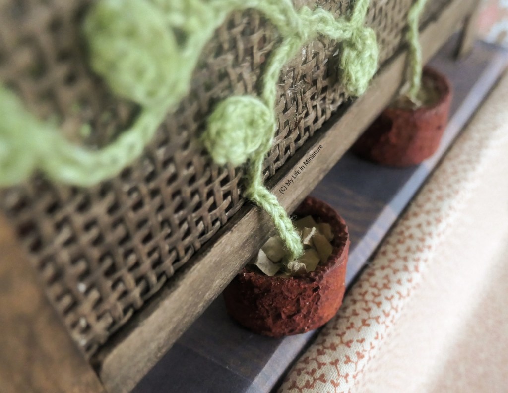 Close-up of one of the pots from above. Inside it the crocheted chain/stem disappears into dirt, or torn-up brown paper. The chain floats over the bottom border of the lattice before attaching to the lattice itself. 