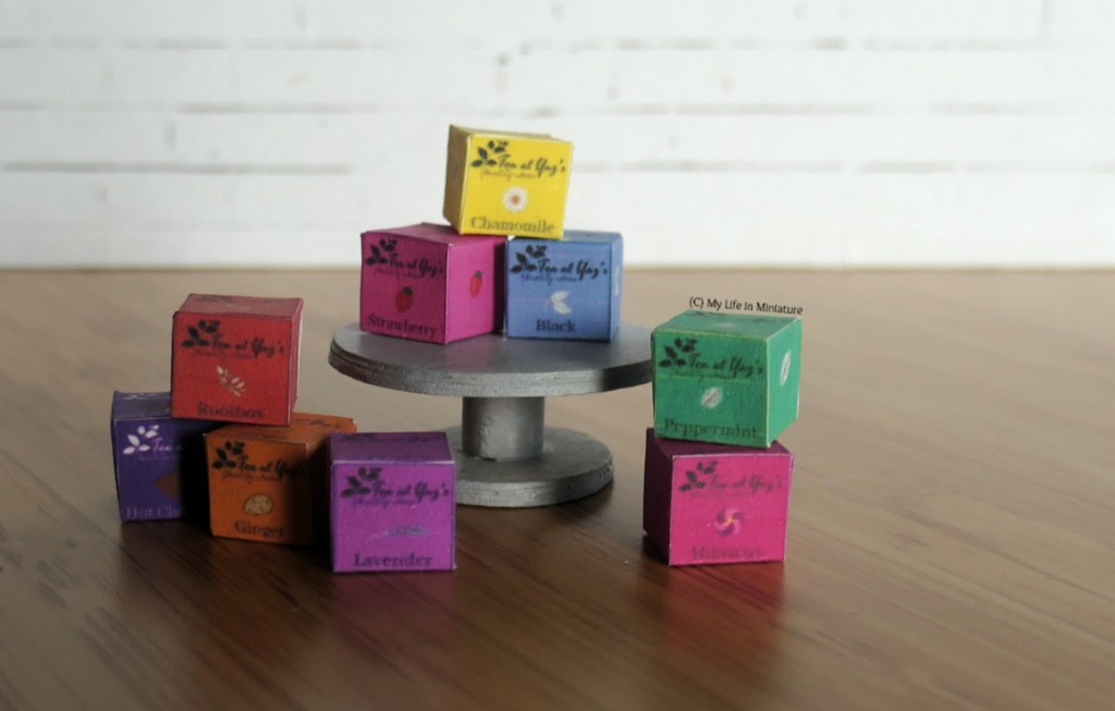 An assortment of Tea at Yaz's tea boxes are arranged around a silver cake stand, against a white brick background. The boxes are in all different colours, and each box is a different tea variety. Visible ones include black, chamomile, peppermint, ginger, and lavender.