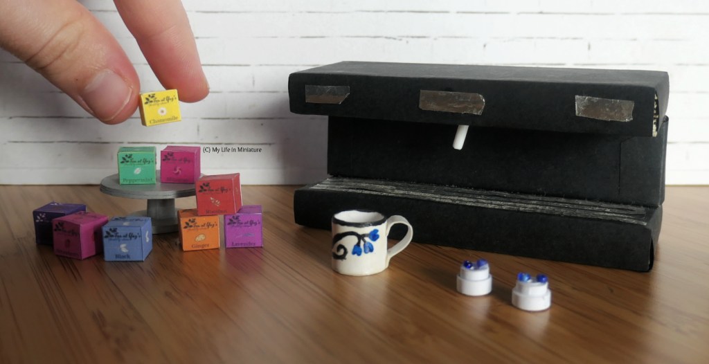 The tea boxes, cake stand, coffee machine, mugs, and coffee machine parts are all photographed together against a white brick background. The author's hand descends from the top left of the image, holding a box of chamomile tea. 