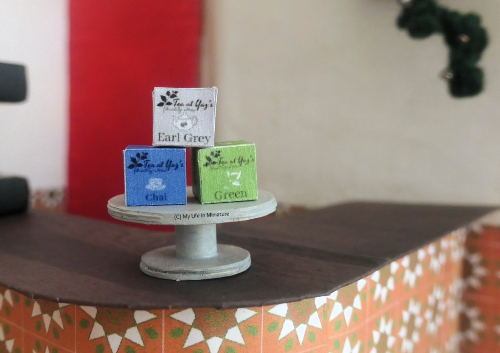 Three Tea at Yaz's tea boxes are arranged in a triangular stack on a silver cake stand on Tea at Yaz's front counter. On top is a grey Earl Grey box, on the bottom left is a blue Chai box, and on the bottom right is a green Green tea box. 