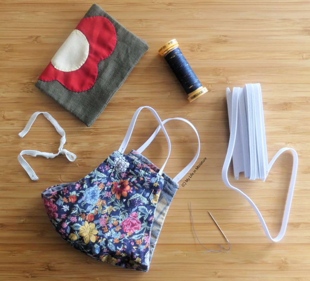 Another arrangment of craft supplies on a wood surface, taken from above. At the centre is a fabric mask with some pins holding new elastic in place. Around the mask is an old, tattered piece of elastic, a needlebook, thread, fresh elastic, and a threaded needle.