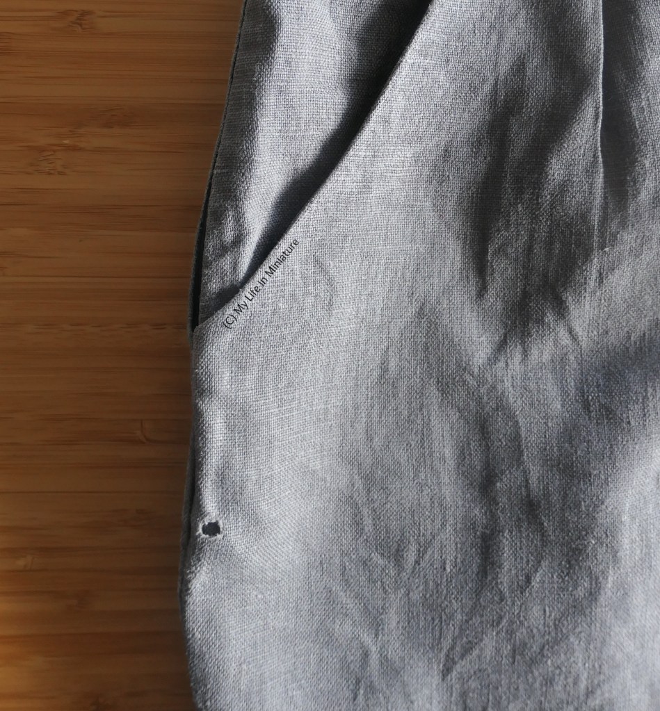 A closeup of the pockets of the author's dark grey pants, laid flat on a wood surface. A small circular hole has opened a small distance below the opening of one of the pockets, close to the side seam. It is quite obvious on the front of the pants.