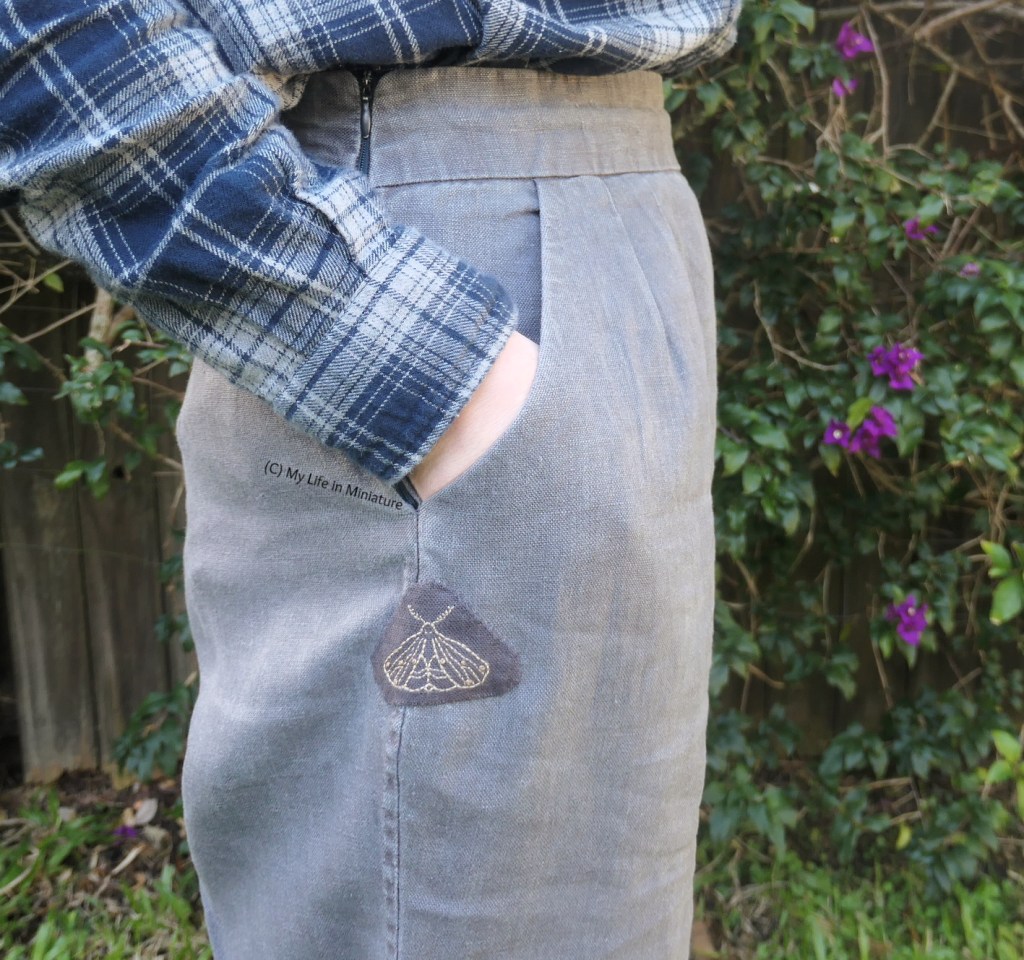 The author stands in front of a hedge, photographed from waist to thigh. She wears dark grey pants with a navy checked shirt. The side seam with the hole faces the camera. Covering the hole is the moth embroidery, stitched onto the pants. The author's hand is in the pocket just above the embroidery.