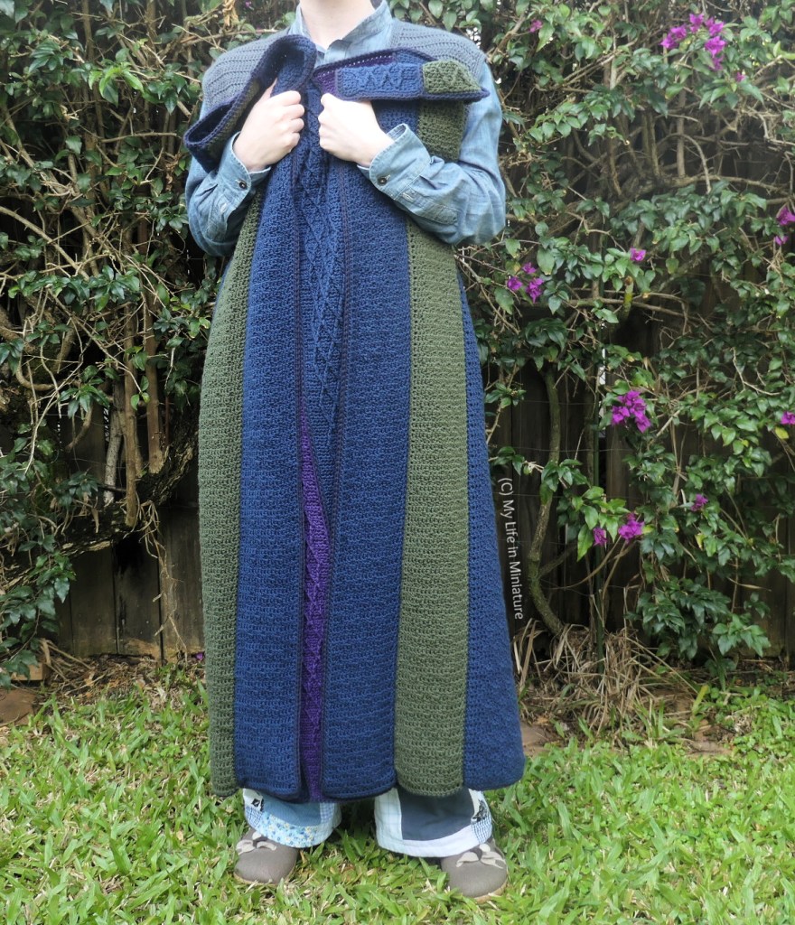 The author stands in front of a hedge, photographed from neck to the ground. She's holding the crochet blanket to her chest, folded over a bit and scrunched up. It falls to just above her ankles, and the back of the crochet strips has no cabling on.