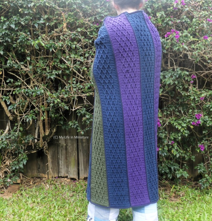 The author stands in front of a hedge, photographed from neck to ankles. She faces away from the camera, with a crocheted blanket wrapped around her. The blanket is made of long strips with a Celtic knotwork design. The strips alternate between navy, purple, and green.