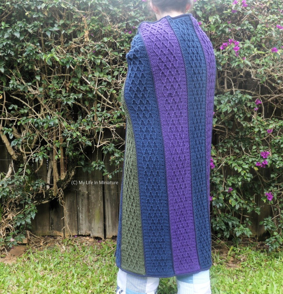 The author stands in front of a hedge, photographed from neck to calves. She stands with her back to the camera, with the crochet blanket wrapped around her back. 
