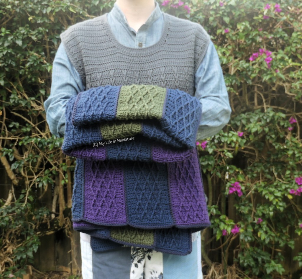 The author stands in front of a hedge, photographed from neck to thighs. She's holding the crochet blanket, folded and thrown over her folded arms. 