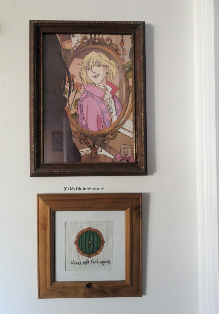 The framed stitching hangs on a white wall, beneath a larger, rectangular frame. The larger frame is dark brown with hints of gold, and has a portrait of Howl from "Howl's Moving Castle" in it.