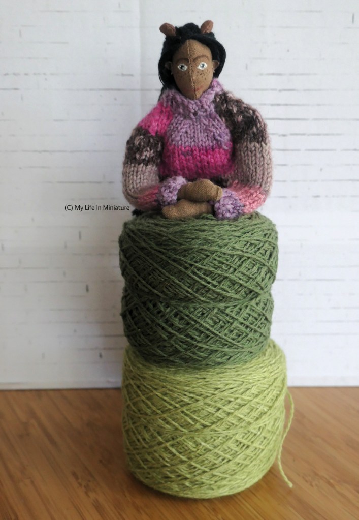 Petra leans on two balls of yarn stacked on top of each other. The top ball is dark green, and the bottom ball is a paler green. She's smiling at the camera, and is wearing a purple and pink knitted jumper.