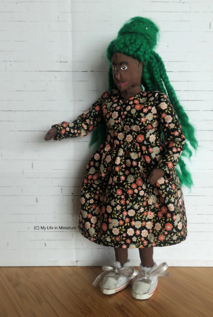 Hazel stands in front of a white brick background, looking down at her outfit. She's wearing a matching set, comprising of a long-sleeved shirt and skirt, made in black fabric with small pink and pale green flowers all over it. She's also wearing white shoes and has half her green braids in a bun.