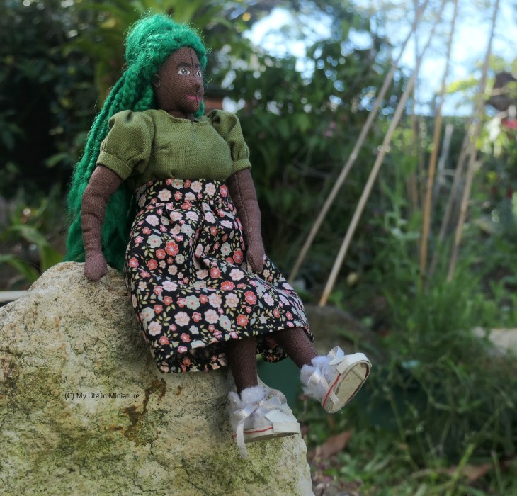 Hazel sits on a rock, legs swinging and one hand braced for balance. She's wearing a black skirt with a pink floral pattern, a green puff-sleeved shirt, and white shoes. She's looking to the right, into the middle distance.