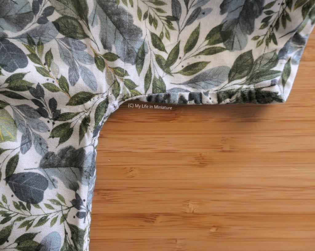 Closeup of the side seam of a short-sleeved, white shirt covered in leaves. There is a small hole fraying at the underarm, where the sleeve turns a corner and becomes the bodice. The shirt is laid on a wood surface.