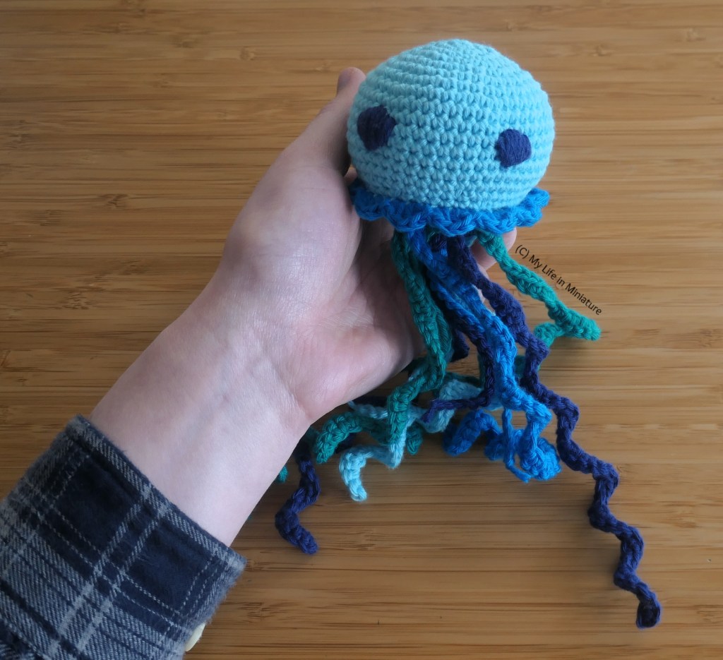The author's hand holds the jellyfish, against a wood background. The body fits in her palm, and the tentacles are significantly longer. Its body is pale blue, with a mid blue frill around the bottom. Its eyes are embroidered in navy blue, and its tentacles are a mix of all the blues and sea green.