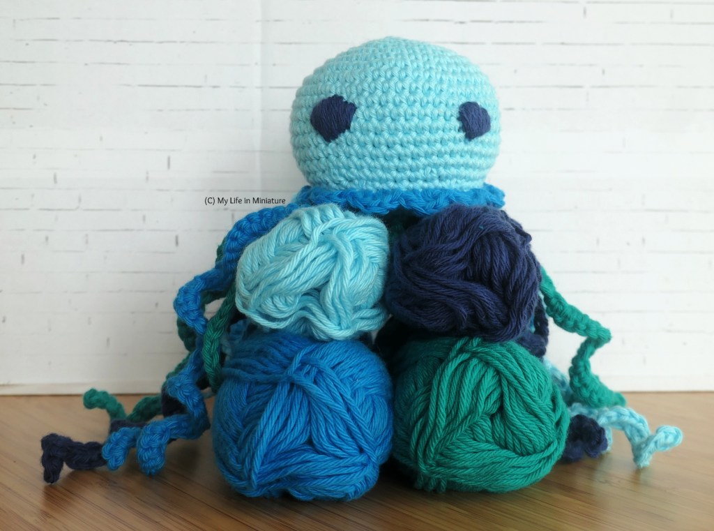 The four colours of yarn used to make the jellyfish are stacked, with the jellyfish seated on top. Its tentacles flow down each side of the stack. Its eyes face the camera, and the background is white brick.