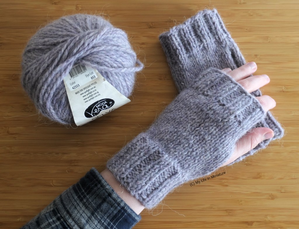 The author's hand, wearing one of the pale grey gloves, rests on top of the other pale grey glove on a wood surface. Beside her hand is the ball of yarn used to make the gloves. 