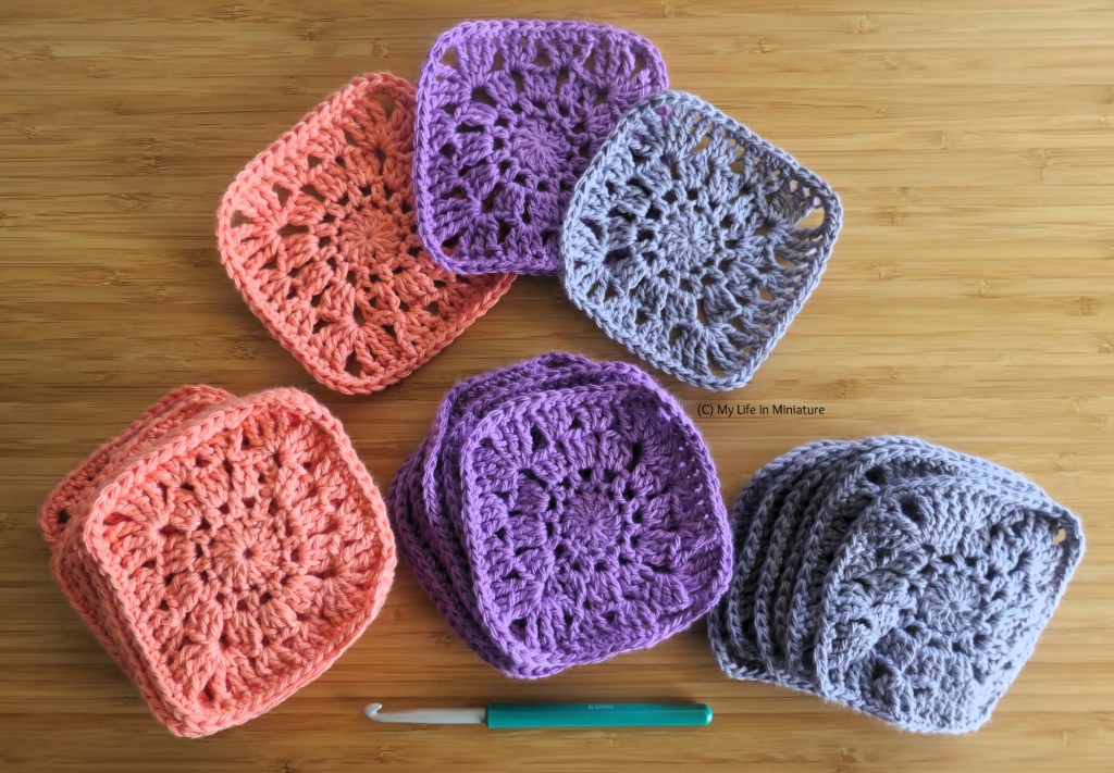 Three crochet granny squares, and three piles of granny squares, are arranged on a wood surface. The top three squares are in peach, purple, and grey, from left to right. The piles corrrespond with these colours, left to right, as well. At the very bottom of the image is a crochet hook.